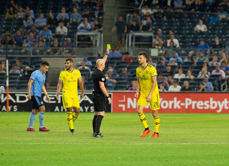 Referee at center issues yellow card to player at right; two other players in background.